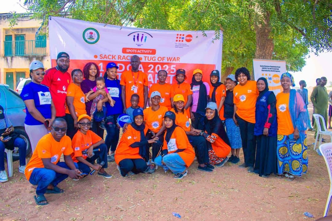 A large group in orange T-shirts and caps posing under a tree before a sports activity banner.